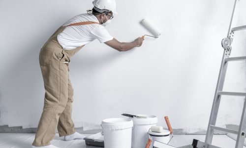 a man paints a white wall with a roller. Repair of the interior. Young male decorator painting a wall in the empty room, concept builder or painter in helmet with paint roller over the empty room.